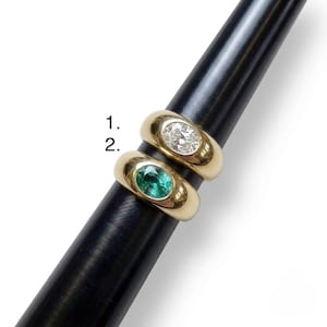 May include: Two gold rings on a black mandrel. Ring 1 features a clear, oval-cut stone. Ring 2 features a green, oval-cut stone. Both rings have a smooth, wide, polished gold band with a bezel setting for the centre stones, labelled 1 and 2.