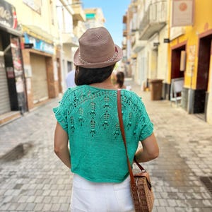 May include: A woman wearing a green crocheted top with a white crocheted trim, white shorts, and a brown woven bag.