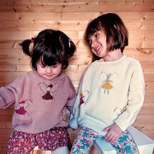 May include: Two young girls wearing knitted sweaters with embroidered fairy designs. The girl on the left is wearing a pink sweater and a floral skirt. The girl on the right is wearing a white sweater and floral leggings.