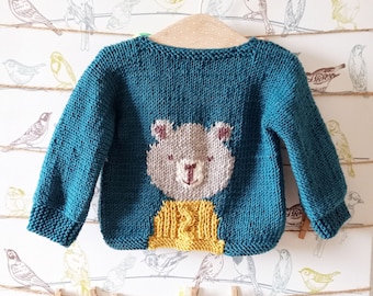 Puede incluir: Un jersey de punto azul turquesa para bebé con una cara de oso amarillo y marrón en la espalda. El jersey está colgado de una percha blanca sobre un fondo blanco con un patrón de pájaros marrones en alambres.