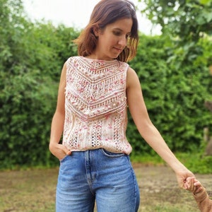 May include: A cream-coloured, sleeveless crochet top with a detailed, geometric pattern in pink and red. The top has a round neckline and is worn with blue denim jeans. The image is set outdoors with a green background.