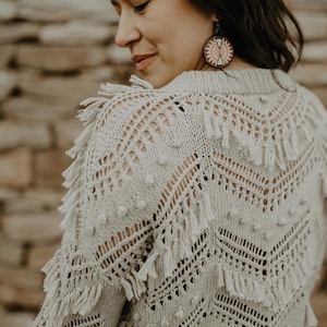 Puede incluir: Un suéter de crochet beige con detalles de flecos y pompones. El suéter tiene un corte holgado y un escote redondo.