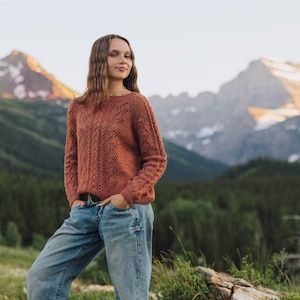 May include: Rust-colored knit sweater with a diamond pattern. The long-sleeved sweater is paired with blue jeans. The background features a mountain range and a forest.