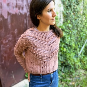 Könnte beinhalten: Ein rosa gehäkelter Pullover mit einem Spitzenmuster und Noppen. Der Pullover hat einen runden Ausschnitt und lange Ärmel.