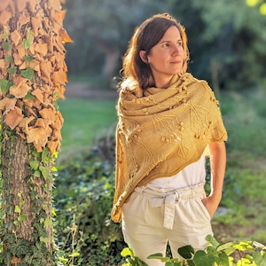 Può includere: Una donna indossa uno scialle di maglia giallo con un motivo a foglie e pantaloni bianchi in piedi in un giardino.