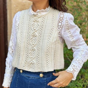 以下が含まれることがあります： クリーム色のケーブル編みと葉模様のニットベスト。ハイネックで、袖にレースのディテールが入った白いブラウスの上に着用。青いデニムジーンズと合わせています。