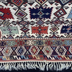 May include: A white rug with a geometric pattern in shades of red, blue, and green. The rug has a fringe border.
