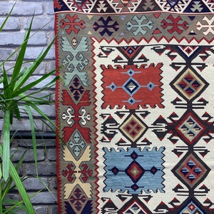 May include: A traditional Turkish kilim rug with a geometric pattern in shades of red, blue, brown, and cream. The rug has a white background and features intricate designs.