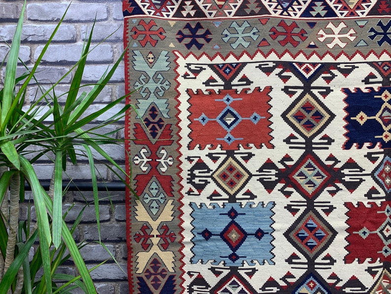 May include: A traditional Turkish kilim rug with a geometric pattern in shades of red, blue, brown, and cream. The rug has a white background and features intricate designs.