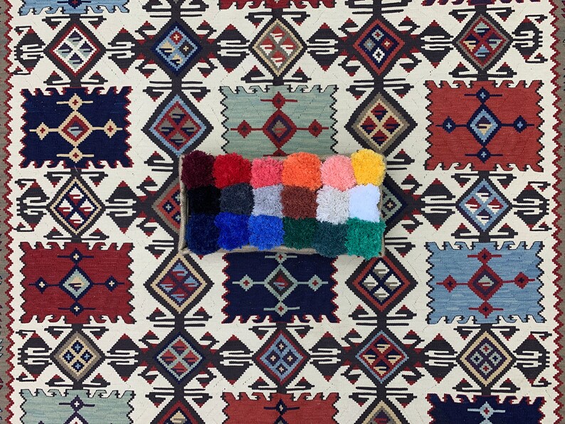May include: A colorful, rectangular pillow with a patchwork design. The pillow is made of different colored fabrics and has a fluffy texture. The pillow is sitting on a white and brown patterned rug.