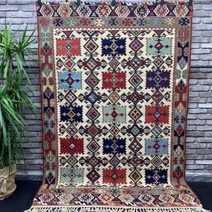 May include: A traditional rug with a geometric pattern in shades of red, blue, green, and brown. The rug has a white background and a fringe border.