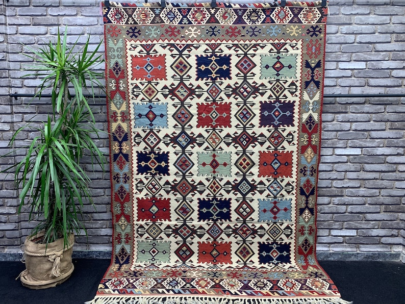 May include: A traditional rug with a geometric pattern in shades of red, blue, green, and brown. The rug has a white background and a fringe border.