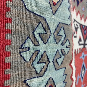 May include: Close-up of a woven rug with a geometric pattern in shades of blue, red, white, and gray. The rug has a fringe border.