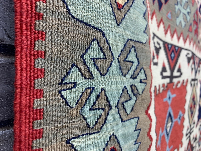 May include: Close-up of a woven rug with a geometric pattern in shades of blue, red, white, and gray. The rug has a fringe border.