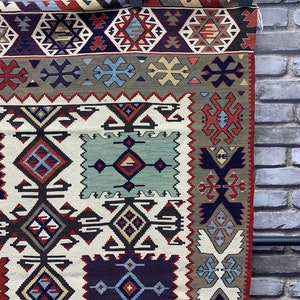 May include: A traditional rug with a white background and intricate geometric patterns in shades of red, blue, and brown. The rug has a fringe border and is woven with a variety of textures.