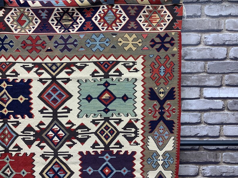 May include: A traditional rug with a white background and intricate geometric patterns in shades of red, blue, and brown. The rug has a fringe border and is woven with a variety of textures.