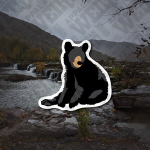 Op de afbeelding: Een sticker met een zwart berenjong met een witte omtrek, zittend op een rotsachtig oppervlak in de buurt van een waterval. De beer heeft een lichtbruine neus en kijkt recht in de camera.