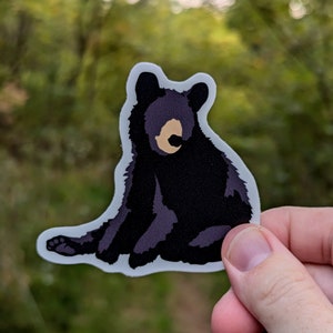 Op de afbeelding: Een sticker met een zwart berenjong met een witte omtrek. De beer zit op zijn achterpoten met zijn poten ingetrokken. De sticker is op een witte achtergrond.
