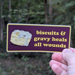 May include: A purple and yellow sticker with a cartoon drawing of a biscuit with white gravy on top. The text on the sticker reads "biscuits & gravy heals all wounds".