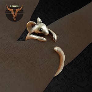 May include: A silver-toned ring shaped like a mouse, with the mouse's head and paws forming the top of the ring. The tail curves around to meet the head. The ring is set against a dark background with a decorative pattern. The word "LEATHER" is in the upper left corner.
