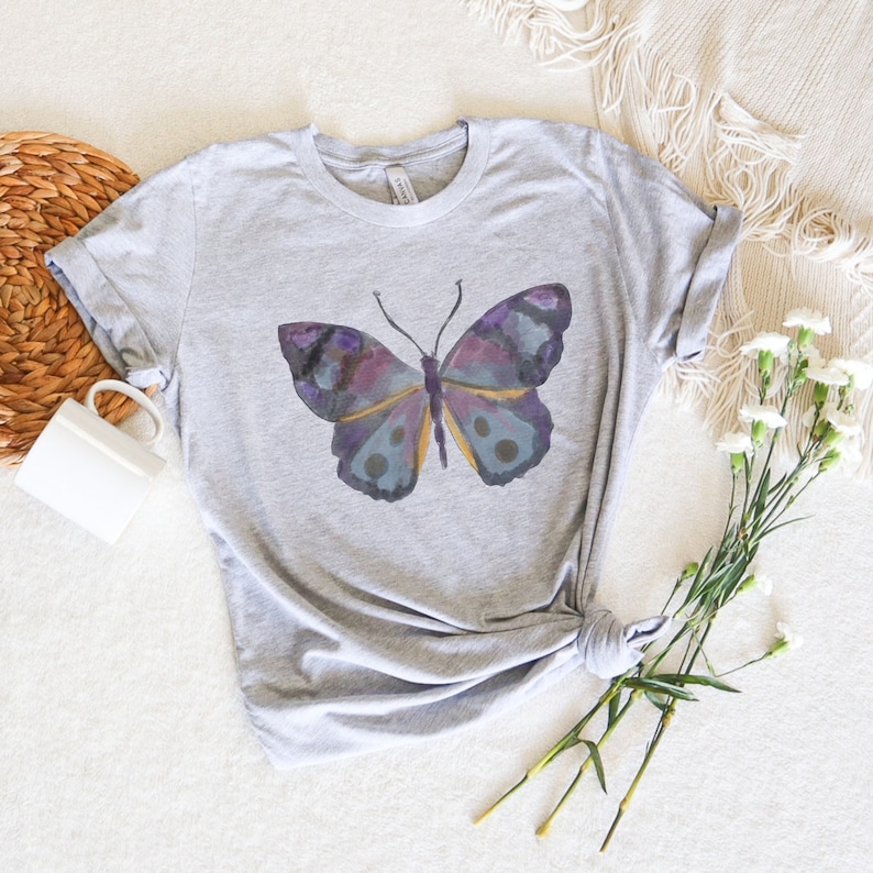 May include: A light gray t-shirt featuring a watercolor butterfly design. The butterfly has purple, blue, and yellow accents. The shirt is knotted at the waist and is displayed on a white surface with a mug and flowers.