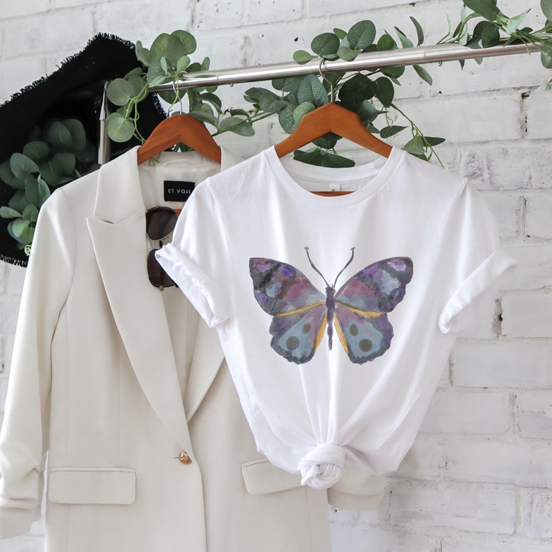 May include: White t-shirt featuring a watercolor butterfly design in shades of purple, blue, and yellow. The shirt is displayed on a wooden hanger alongside a cream-colored blazer and sunglasses. The overall aesthetic is clean and modern.