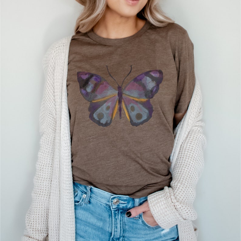 May include: Brown t-shirt featuring a watercolor butterfly design. The butterfly has purple, blue, and yellow accents. The shirt is paired with blue jeans and a cream-colored cardigan.