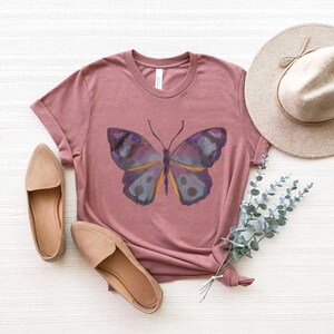 May include: A mauve-colored t-shirt featuring a watercolor butterfly design in shades of purple, blue, and yellow. The shirt is styled with a pair of tan flats and a wide-brimmed hat, creating a casual, feminine aesthetic.