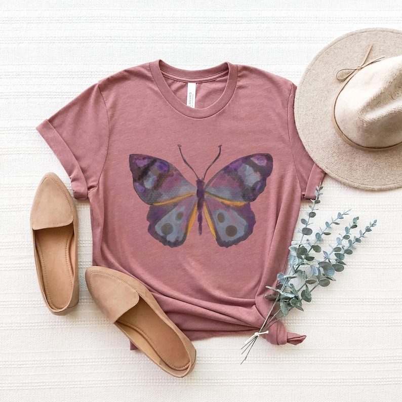 May include: A mauve-colored t-shirt featuring a watercolor butterfly design in shades of purple, blue, and yellow. The shirt is styled with a pair of tan flats and a wide-brimmed hat, creating a casual, feminine aesthetic.
