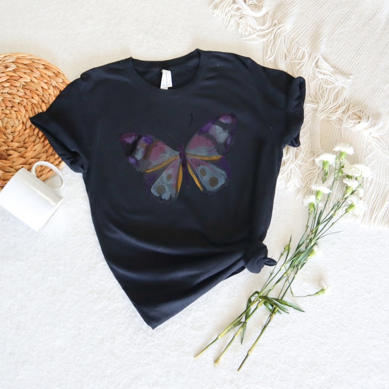 May include: Black t-shirt featuring a colorful butterfly design. The butterfly has purple, blue, and yellow accents. The shirt is folded and knotted at the bottom. White flowers and a woven basket are in the background.