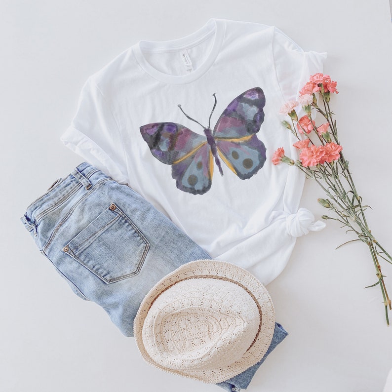 May include: White t-shirt with a watercolor butterfly design in shades of purple, blue, and yellow. The shirt is paired with light blue denim jeans and a straw hat. Pink flowers are arranged to the side, creating a casual, summery aesthetic.
