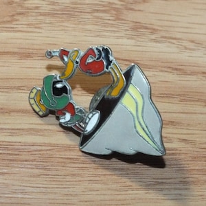 May include: A metal pin featuring a cartoon image of Marvin the Martian and Tweety Bird. Marvin is green and red, and Tweety is yellow and red. The pin is on a wooden surface.