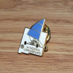 May include: A gold-colored pin with a white background and a blue sailboat. The text "WINDSAIL LORDSBURG" is printed on the pin.