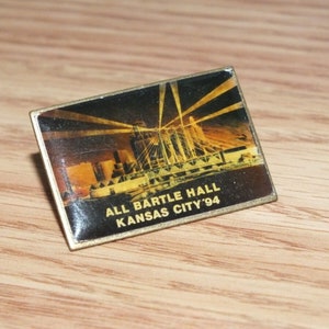 Op de afbeelding: Een gouden pin met een zwart-wit afbeelding van een stadsbeeld 's nachts. De tekst "ALL BARTLE HALL KANSAS CITY '94" staat op de pin gedrukt.