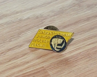 Puede incluir: Un pin de esmalte amarillo y azul con el texto "Saskatchewan Junior Hockey League" y la abreviatura "SJHL". El pin presenta un palo de hockey y un disco estilizados.