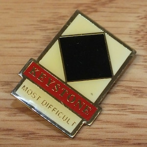Op de afbeelding: Een gouden en rode emaille pin met de woorden "Keystone Most Difficult" en een zwarte diamantvorm in het midden.
