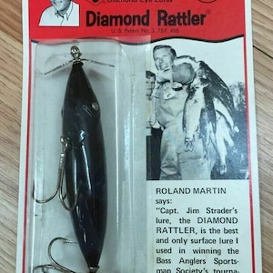 May include: A black and silver fishing lure called "Diamond Rattler" is packaged in a clear plastic blister pack. The packaging features a red background with white text and a picture of a man holding a fish. The text on the packaging reads "Capt. Jim Strader's Diamond Eye Lures Diamond Rattler U.S. Patent No. 3,757,455". The text below the picture of the man reads "Roland Martin says: "Capt. Jim Strader's lure, the DIAMOND RATTLER, is the best and only surface lure I used in winning the Bass Anglers Sportsman Society's tournament on Lake Seminole, Georgia."
