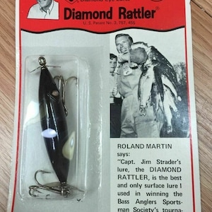 May include: A black and green fishing lure with a white eye and a diamond-shaped design. The lure is packaged in a plastic blister pack with a red and white card that reads "Capt. Jim Strader's Diamond Eye Lures Diamond Rattler® U.S. Patent No. 3, 757, 455". The card also features a photo of a man holding a string of fish and the text "Roland Martin says: "Capt. Jim Strader's lure, the DIAMOND RATTLER, is the best and only surface lure I used in winning the Bass Anglers Sportsman Society's tournament on Lake Seminole, Georgia."
