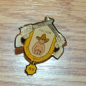 May include: A gold-colored pin with a cartoon cowboy baby wearing a hat and holding two pistols. The pin says "Mac's Mavericks" and "Tucson" with the number "88" on the bottom.