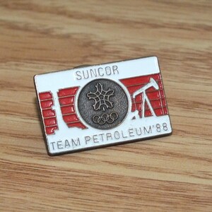 Op de afbeelding: Een witte en rode emaille pin met de tekst "SUNCOR TEAM PETROLEUM '88". De pin heeft een gestileerde olieboring en de Olympische ringen.