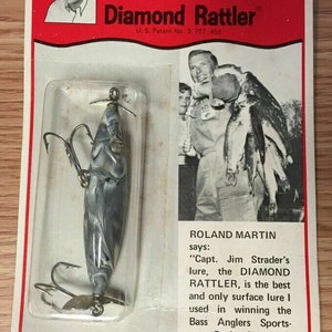 May include: A fishing lure with a white and blue marbled finish, two treble hooks, and a diamond-shaped eye. The lure is packaged on a red and white card with the text "Diamond Rattler" and "Capt. Jim Strader's Diamond Eye Lures".