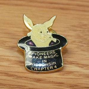 May include: A gold-colored pin with a black top hat and a white rabbit peeking out. The hat has the text "Pioneers Make Magic in Wisconsin Chapter 4".