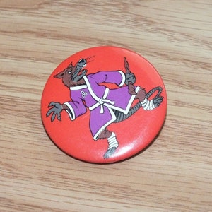 Puede incluir: Un botón rojo con una imagen de dibujos animados de una rata marrón que lleva un kimono de karate morado con un cinturón blanco. La rata está corriendo y tiene un vendaje blanco en el tobillo derecho. La rata tiene un símbolo blanco en el pecho que parece un círculo con tres círculos más pequeños dentro.