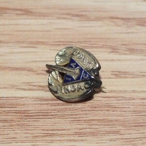 Pode incluir: Um alfinete dourado com um triângulo de esmalte azul com as letras "I.A.C." e as palavras "Junior" e "Indac".