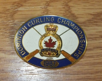 Puede incluir: Pin ovalado de los Campeonatos de Curling Dominion con borde azul y letras doradas. El centro presenta un fondo blanco con una hoja de arce roja y el texto "LEGION" y "1987". Dos escobas de curling en la parte inferior.