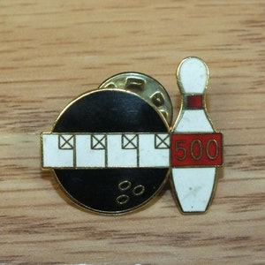 May include: A gold-tone bowling pin and ball pinback button. The bowling pin is white with a red stripe and the number 500 on it. The ball is black with white pins and three black bowling balls.