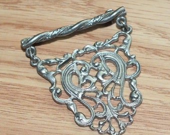 May include: A silver filigree pendant with a twisted metal bar attached. The pendant is in the shape of a heart with intricate details.