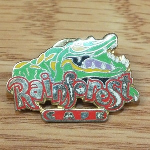 Puede incluir: Un pin de color dorado con una cabeza de cocodrilo verde, amarilla y negra y las palabras "Rainforest Cafe" en letras rojas y blancas.