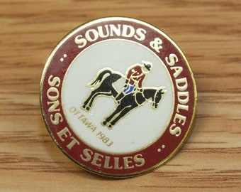 Puede incluir: Un pin de color dorado con un diseño circular rojo y blanco. El pin presenta un caballo negro con un jinete con un uniforme rojo y azul. El texto "SOUNDS & SADDLES SONS ET SELLES" está escrito en un círculo alrededor del caballo y el jinete. El texto "OTTAWA 1983" está escrito debajo del caballo.