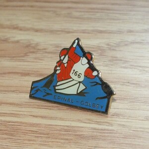 May include: A gold-colored pin with a blue and white image of a canoe with two people paddling. The canoe is numbered 166 and the text "EPINAL - GOLBEY" is below the canoe.